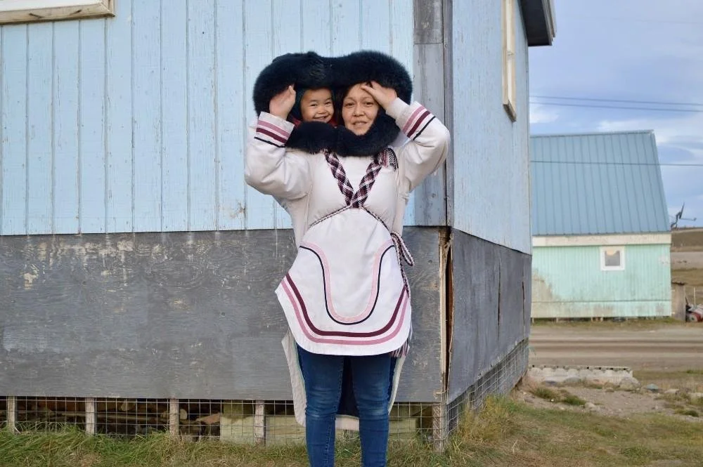 Standing outside a home in the far north, a mother beams as her child peeks joyfully over her shoulders, nestled safely inside a traditional amauti. This amauti, made from white cloth with deep maroon trim and a wide fur-lined hood, reflects a contem