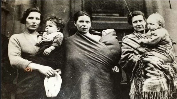 Three Scottish women stand outside a tenement doorway, each with a child close — one carried in arms, one wrapped in wool, one settled across the hip. The photograph, likely taken in Glasgow or another urban centre in the early 20th century, is an ex