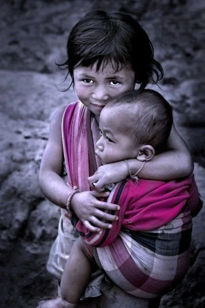 A young girl smiles shyly at the camera as she holds a baby on her hip, wrapped snugly in a checked cloth that loops over her shoulder and under the child’s bottom. The simple, secure carry — likely improvised from a traditional scarf or sarong — dem