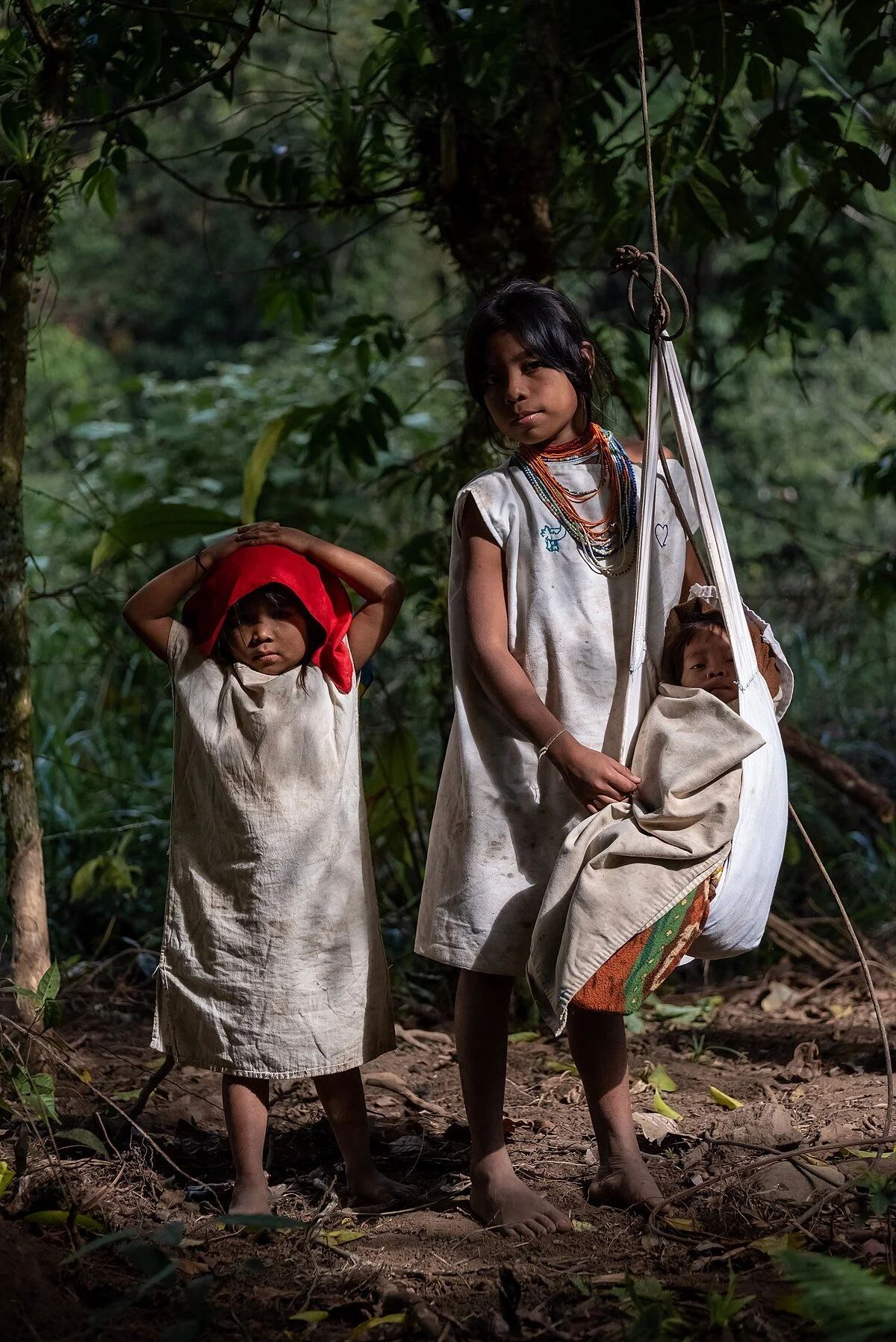 A Kogi mother and daughter stand barefoot along the moss-covered paths of Colombia’s Sierra Nevada, dressed in their distinctive handspun cotton garments. The woman holds a beaded piece of weaving-in-progress — perhaps a small mochila, the iconic bag