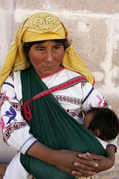 A woman sits against a stone wall, breastfeeding her baby who is carried snugly in a deep green woven rebozo. The rebozo crosses diagonally over her shoulder and supports the baby in a secure front carry. The child’s head is cradled close to her ches