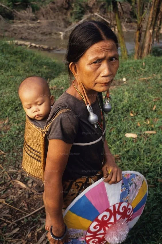 A mother from the highland regions of northern Thailand or Laos carries her sleeping child in a woven basket carrier, a style typical of many Indigenous hill tribes in Southeast Asia. The child is nestled securely in the upright carrier, their body g