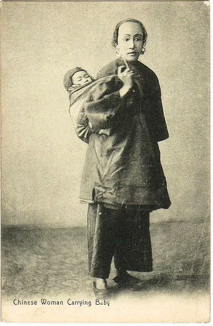 A quiet studio portrait captures a Chinese woman carrying her baby in a traditional back-carry, most likely with a rectangular bei dai or similar wrap cloth. Her baby rests high and secure against her back, arms tucked close, the fabric gathered snug