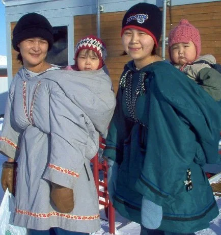 Two Inuit mothers stand side by side in the snow, each carrying a child tucked warmly into the back of a traditional amauti. These modern amauti garments, sewn from cloth and insulated for Arctic conditions, maintain the essential design that has sup