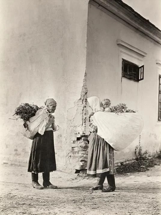 This photograph, likely taken in Eastern Europe in the early 20th century, shows two rural women standing beside a whitewashed building. Both are wearing long skirts, aprons, headscarves, and practical shoes typical of traditional Eastern European pe