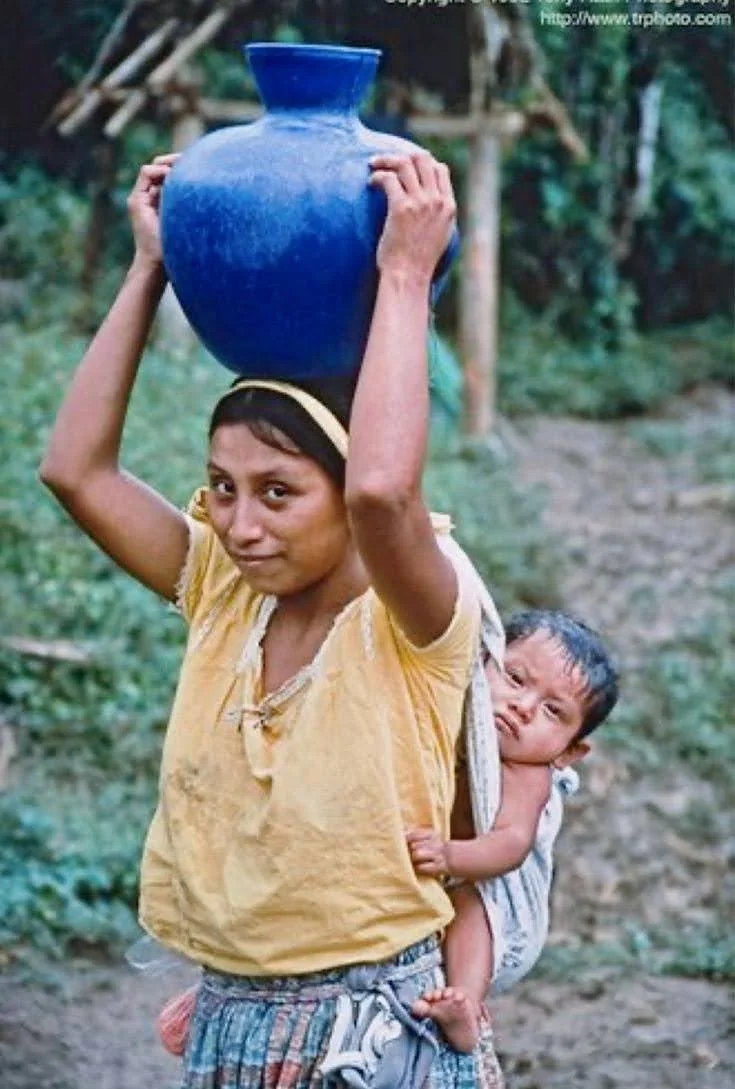 This vivid photograph captures a young Indigenous Guatemalan mother from a rural community, effortlessly balancing a large blue ceramic vessel on her head while carrying her baby on her back. The child is secured with a traditional rebozo-style cloth