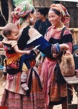 Two women of the Flower Hmong community share a moment in a bustling village market in northern Vietnam. Dressed in the vivid, richly embroidered skirts, aprons, and headscarves that distinguish their group, one carries her baby on her back using a c