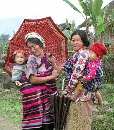 In a lush rural village in Myanmar, two women pause for conversation with babies carried securely on their backs. Each baby is wrapped in a check-patterned cloth, tied across the mother’s chest in a wide band that supports the child’s weight and ensu