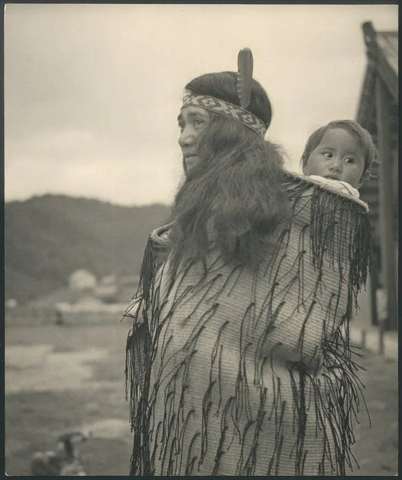 Standing tall against the backdrop of the whenua, a Māori mother wears a finely woven cloak adorned with tāniko borders and black tasselled detailing, carrying her child tucked warmly into the folds at her back. Her long hair falls freely, crowned by