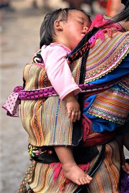 A small child sleeps deeply against her mother’s back, cradled in layers of rich embroidery and handwoven cloth. This is the distinctive style of the Flower Hmong — a subgroup of the Hmong ethnic minority in northern Vietnam — whose carriers are ofte
