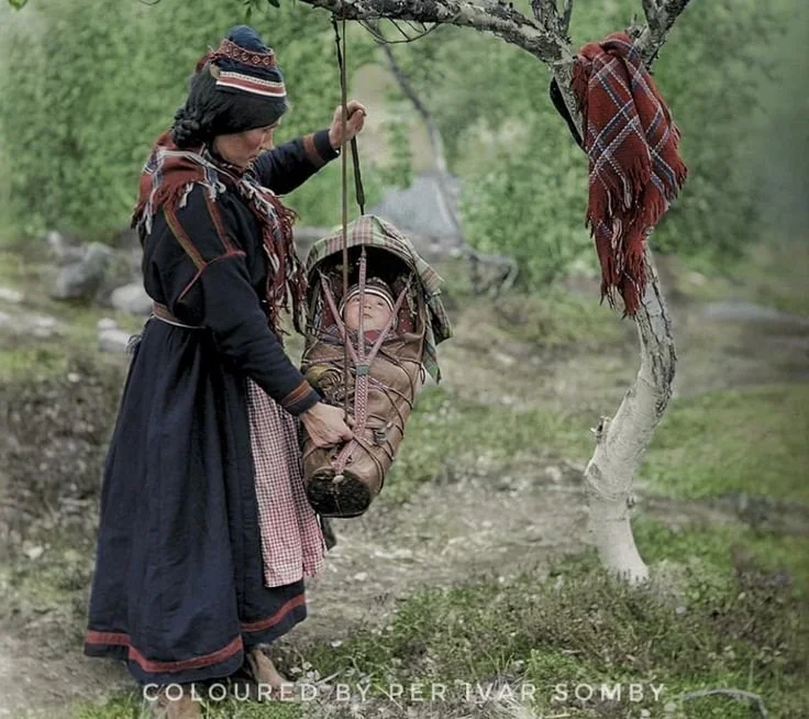 In the northern reaches of Sápmi, a Sámi mother gently steadies her baby’s komse — a traditional cradleboard — as it hangs from the branch of a birch tree. The infant is swaddled securely within the carved wooden carrier, protected by laced leather b