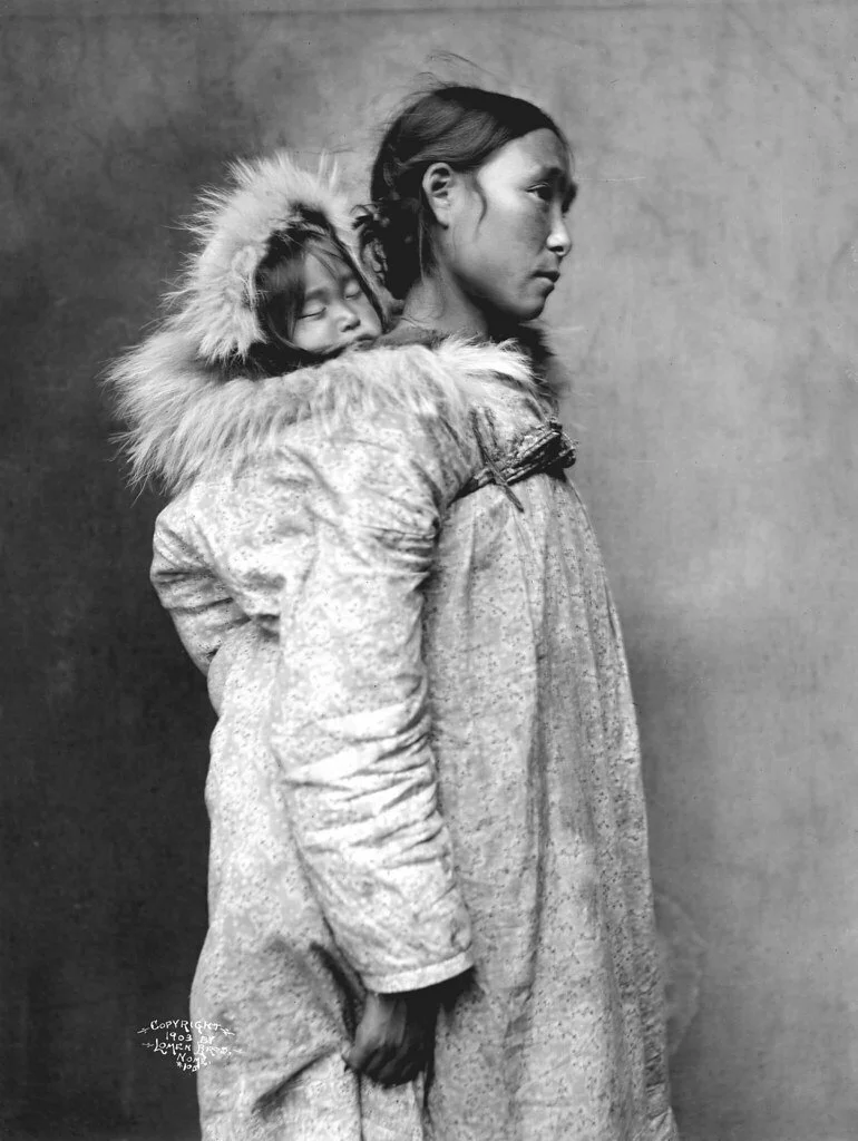In this quiet and powerful 1929 portrait by Edward S. Curtis, an Inupiat (Alaskan Inuit) mother carries her sleeping child nestled deep in the back of her traditional parka. The child's face is framed by a soft halo of fur from the coat’s hood, eyes