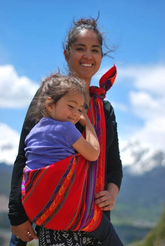Under a sky bright with Andean light, a mother holds her child in a woven embrace. The red manta across her shoulder shimmers with traditional patterns, its stripes echoing the textiles passed down through generations. Though her clothing is modern,