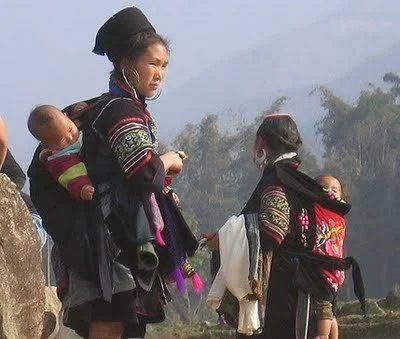 Two Hmong mothers stand outdoors, each carrying a baby high on her back in a richly decorated cloth carrier. The babies are upright and close, with their legs tucked in and their heads supported by the embroidered panels. The carriers are secured wit