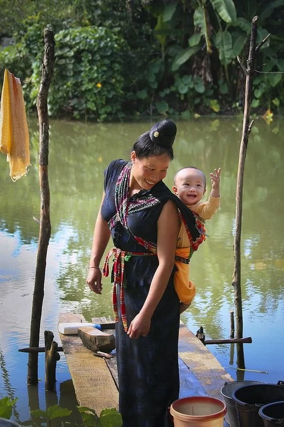 Standing on a narrow bamboo jetty over still, green water, a woman smiles brightly as she carries her baby high on her back. The baby, also beaming, is snugly supported in a soft cloth carrier tied across the mother's chest and around her waist. The