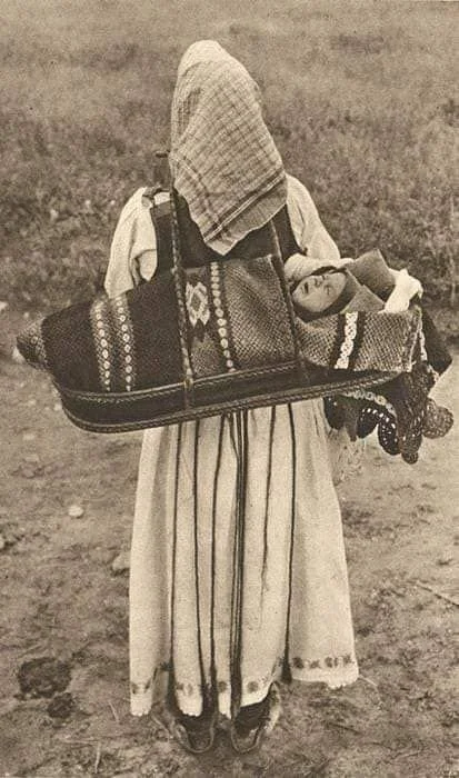A woman walks across the open earth, her head and shoulders covered, her long skirt embroidered with care. Slung over her back is a striking cradleboard — wide, flat, and woven with bold geometric patterns — holding a baby swaddled in thick blankets.
