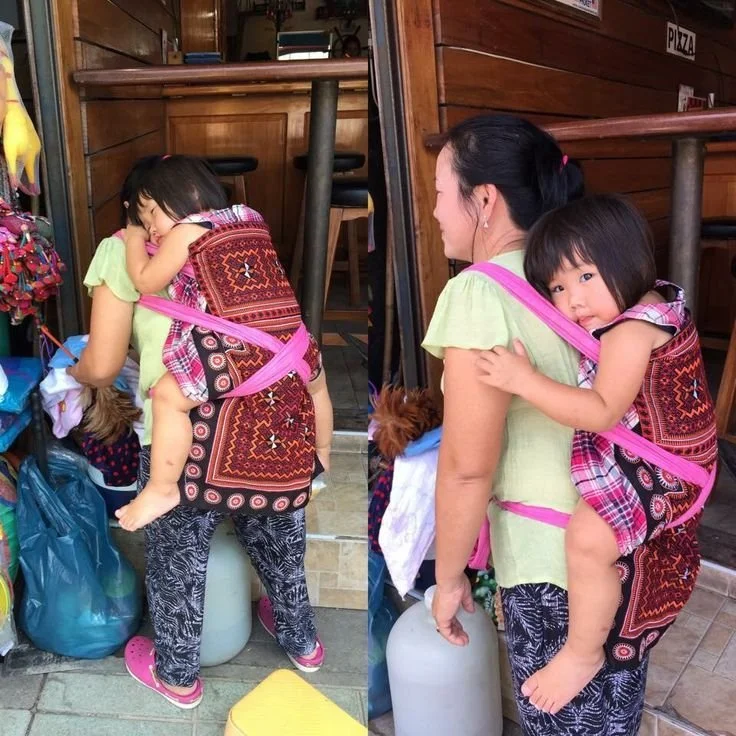 A little girl rests contentedly against her mother’s back, carried in a richly embroidered panel carrier secured with contrasting pink shoulder straps. The traditional textile features bold geometric patterns and circular motifs, stitched with care a