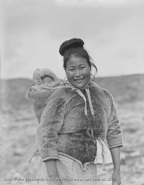 With a radiant smile and upright stance, this Inuit mother carries her baby in a soft back-carry, wrapped securely in a fur-lined garment adapted for Arctic life. Unlike the amauti — which encloses the baby in a built-in parka pouch — this style feat