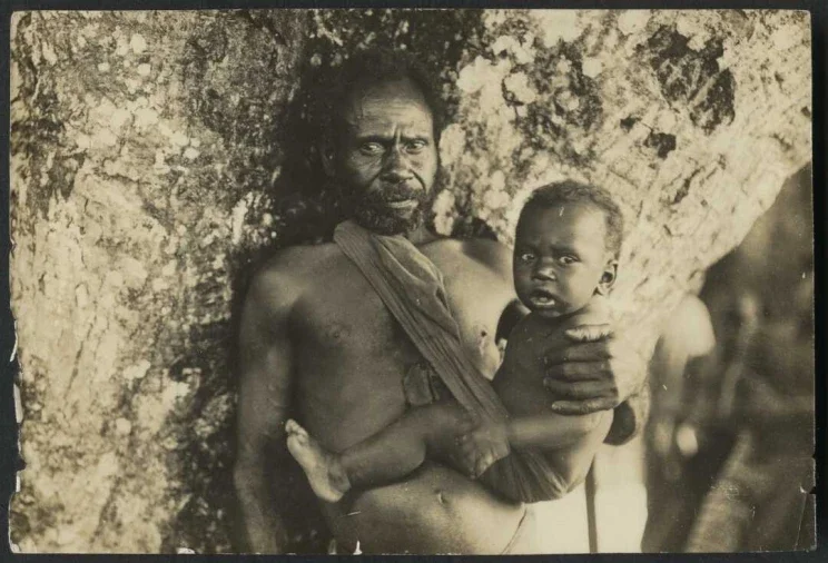 A father stands beneath the dappled bark of a tree, his chest bare and posture steady as he holds his child close. A narrow cloth is looped over one shoulder, securing the baby against his side in a style both instinctive and deeply rooted in traditi