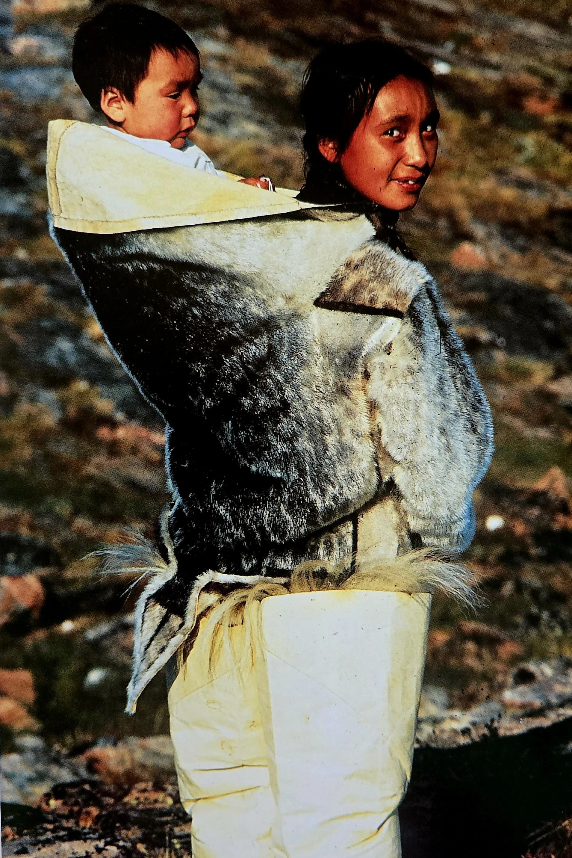 In this striking image from Greenland, a young child is carried close against their mother’s back, nestled inside the traditional amauti — a warm, fur-lined parka designed specifically for carrying infants. The child’s head peeks out from the wide op