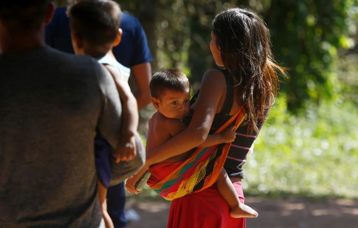 A young mother walks in dappled light, her baby resting securely in a brightly woven cloth slung low around her hip. The child’s eyes meet the camera, wide and watchful, while one arm wraps naturally around their mother’s side. The sling — simple yet
