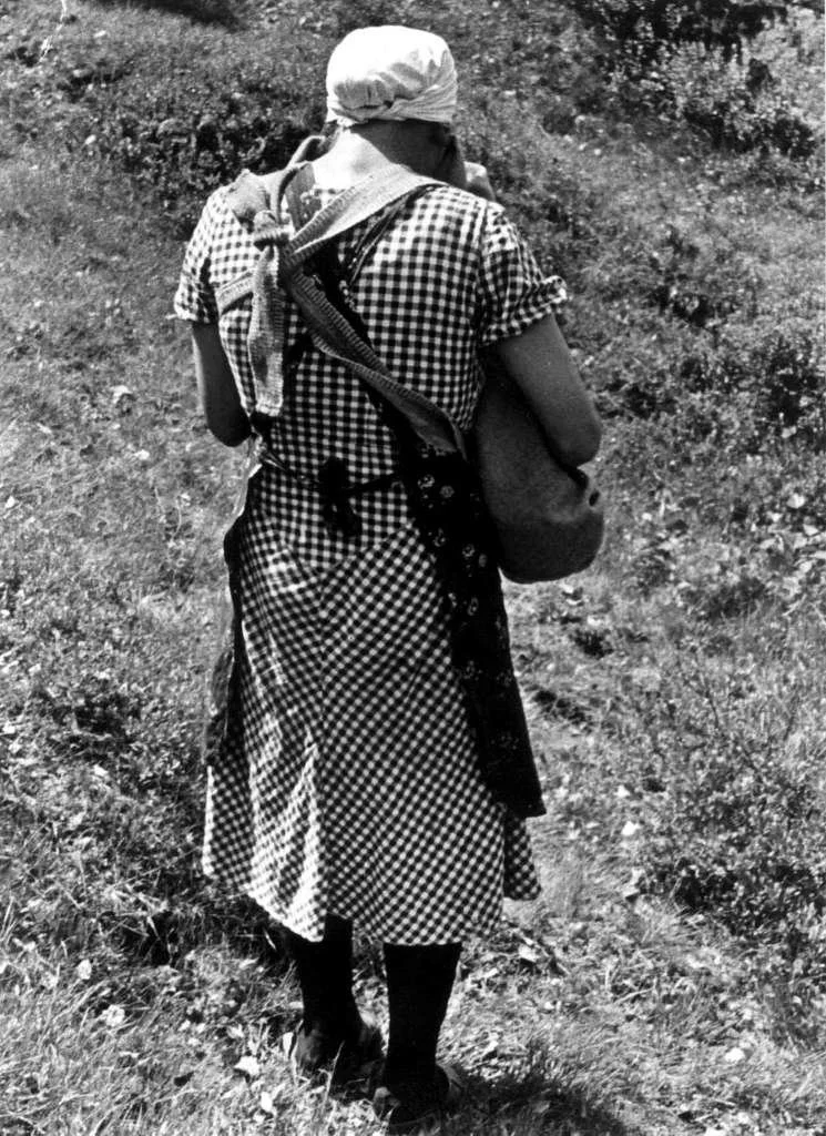 Climbing a grassy slope in southern Norway in 1941, this mother carries her baby tucked safely in a linde, a traditional soft carrier tied at the front of her body. Photographed in Engedal, Fjotland, in the municipality of Kvinesdal, she wears a chec