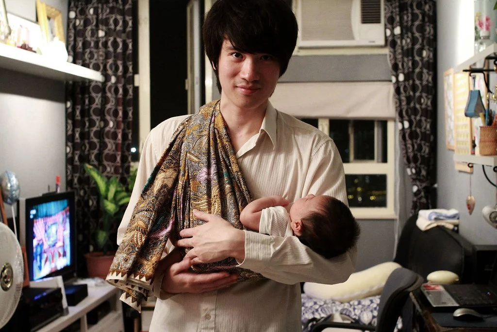 A father stands calmly in the glow of a softly lit room, holding his newborn close in a traditional batik-patterned sling. The cloth, likely a selendang or sarong, is draped over one shoulder and gathered at the hip, cradling the baby securely in an