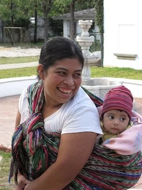 A woman stands smiling broadly in an outdoor courtyard, her baby securely wrapped to her back in a long, multicoloured woven cloth. The fabric is richly textured with streaks of green, red, black, and purple, looped and tied across both shoulders in
