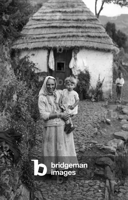A woman stands outside a stone cottage with a thatched roof, cradling a toddler on her hip. Her head is wrapped in a plain scarf, and her clothing is practical and worn, suited to a life shaped by the land. Though not using a carrier in this moment,