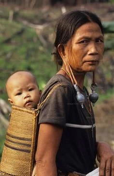 A woven cradle on the back, a sleeping baby within, and the river not far behind — this image holds the quiet strength of tradition. The woman stands tall on the grassy riverbank, a bold fan in her hand and heavy silver ear ornaments framing her face