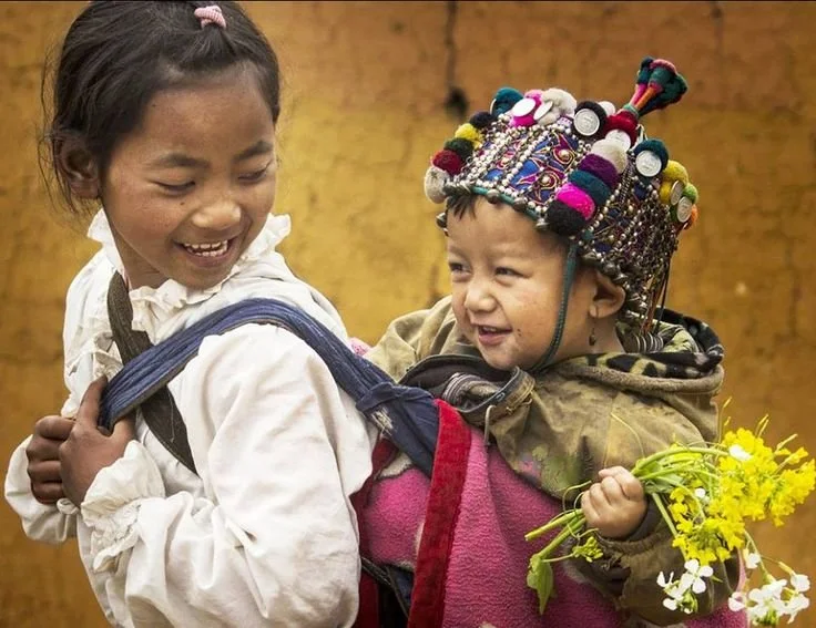 A young girl carries her sibling with ease and joy, both children sharing a moment of laughter in the warm light of their surroundings. The carrier is a simple but effective cloth wrap, likely a traditional mei tai or similar rectangular carrier, tie