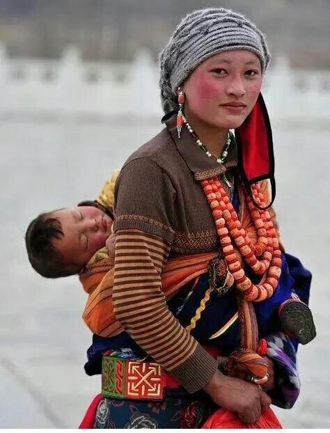 A woman of Sikkimese ethnicity, in the far northeastern reaches of India near the borders of Nepal and Bhutan, beams with joy as she stands in the doorway of a modest metal-walled home. On her back, a young child peeks out from a cheerful patchwork q