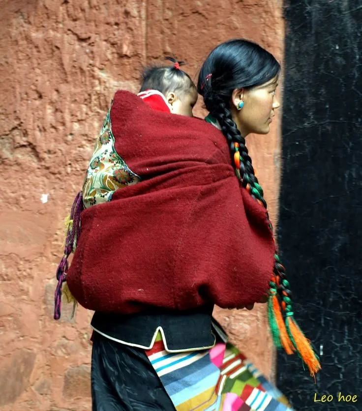 In a village nestled within the hills of northern Laos, a mother carries her young child on her back using a traditional striped carrier cloth, intricately woven in green with accents of deep pink and black. The baby is snug against her body, support