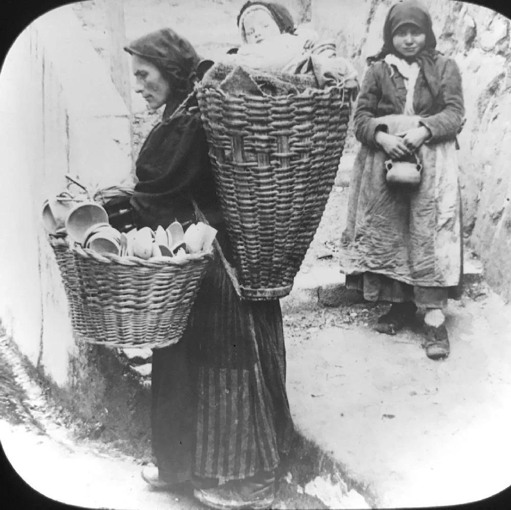 High in the rugged mountains of Cantabria, another Pasiega woman bears the weight of work and motherhood with quiet endurance. Balanced across her back is a tall, tightly woven basket—cradling a child among folded cloth and straw. A second, wide bask