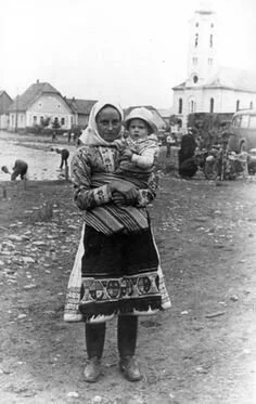 In the heart of an Eastern European village, a mother stands proudly in full traditional dress, her baby held snug against her hip with a finely woven cloth. Her apron and skirt are richly embroidered, with patterns echoing regional folk motifs. A he