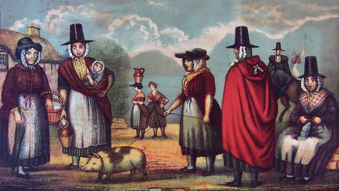 A richly coloured illustration from 19th-century Wales, this scene captures a lively gathering of women in traditional Welsh costume — tall black stovepipe hats, patterned shawls, woven aprons, and woollen cloaks. At the heart of the image stands a m