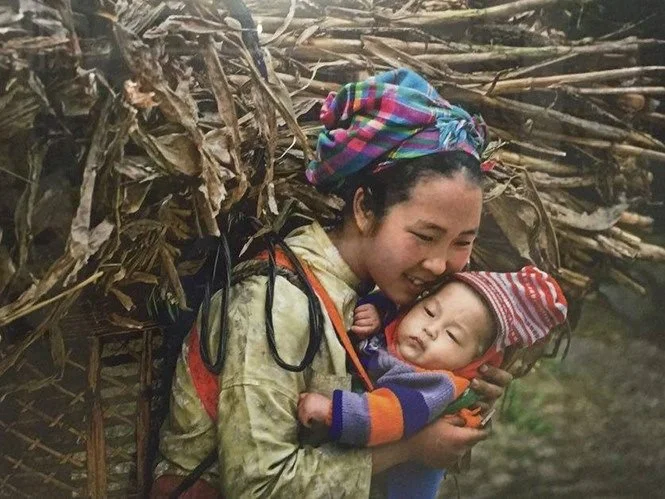 A young mother smiles gently at her baby, who is nestled securely on her back in a traditional carrier. Her arms wrap around the child in a moment of tender affection, even as she bears a heavy load of gathered firewood — a striking image of strength