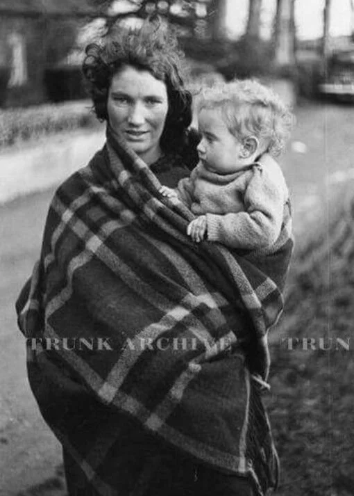 This striking image captures a mother in Britain in the mid-20th century, her child securely wrapped against her in a large checked wool blanket. The cloth is drawn high over her shoulder and around the toddler’s back, supporting the child in a hip c