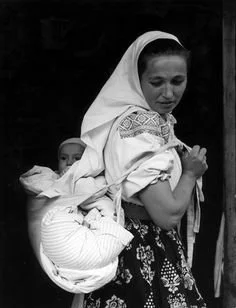 A woman dressed in a traditional embroidered blouse and patterned skirt carries her baby in a cloth carrier tied securely across her back. Her headscarf and the style of both embroidery and wrap suggest a Central or Eastern European setting — most li