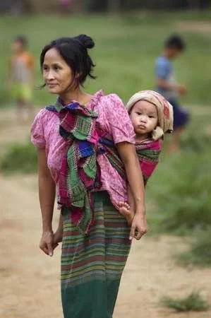A Hmong woman walks through a bustling market, her child carried securely against her back in a bold, floral-printed cloth. The vibrant red fabric, tied over her indigo-dyed clothing, contrasts beautifully with the intricate hand-embroidered bands on