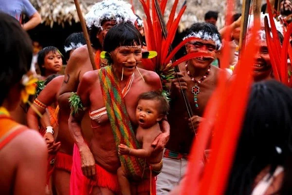 Amid vibrant red feather adornments and celebratory movement, a mother carries her baby close during a cultural gathering of the Kayapo people of Brazil. Her handmade carrying cloth, rich with green and ochre stripes, is draped diagonally across her