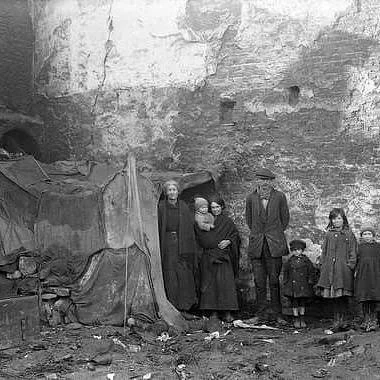 In front of a crumbling plaster wall, a family stands solemnly, gathered close outside what appears to be a makeshift shelter assembled from tarpaulins, cloth, and salvaged boards. The ground beneath them is littered with debris. Their clothing is la