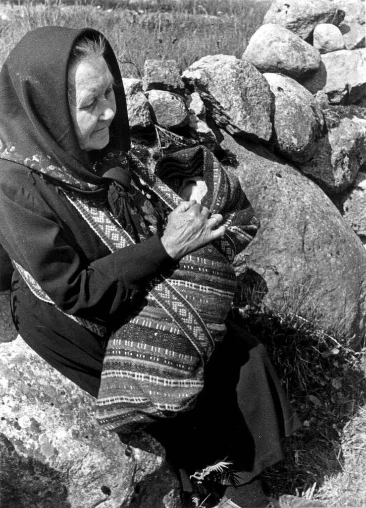 Seated on a sun-warmed rock beside a dry-stone wall, an older woman cradles a baby wrapped snugly in a richly patterned woollen shawl. The bundle is held in the traditional linde style — a way of wrapping that has been used in parts of southern Norwa