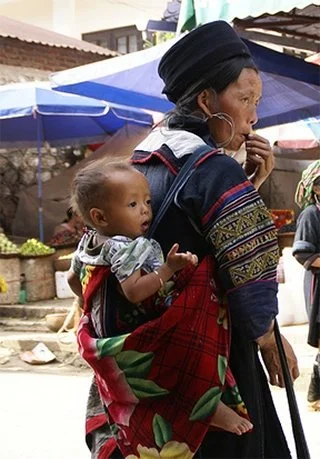 A Hmong woman walks through a bustling market, her child carried securely against her back in a bold, floral-printed cloth. The vibrant red fabric, tied over her indigo-dyed clothing, contrasts beautifully with the intricate hand-embroidered bands on