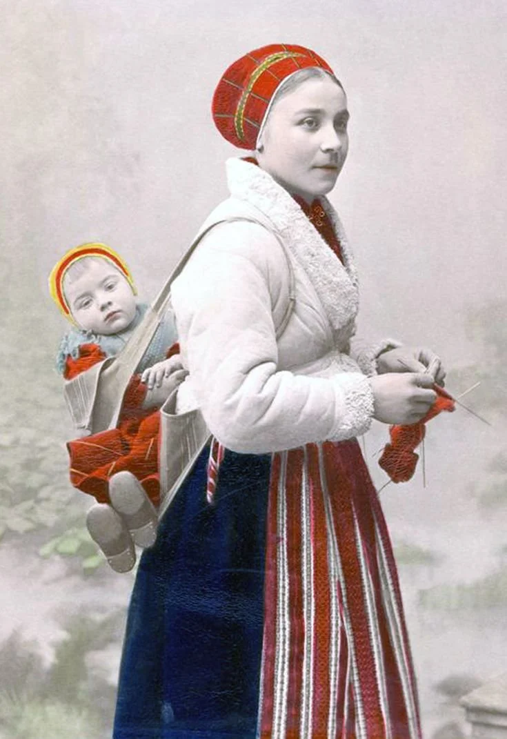 A young Swedish mother carries her baby high on her back, the child securely wrapped in a wide woven sash tied diagonally across her chest — a traditional method seen in many Scandinavian regions. The mother’s hands are busy with her knitting, a comm