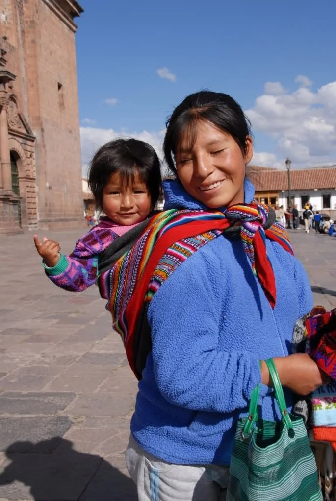 In the bright plaza light of Cusco, a mother walks with her daughter tied securely to her back in a handwoven manta. Their colours sing against the stone streets — red, green, purple, and gold — wrapping them in a tapestry of belonging. The little gi