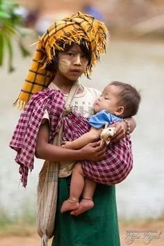 This striking image captures a young girl in Myanmar (Burma) gently carrying her baby sibling in a simple cloth sling. The cloth, likely a longyi or similar traditional garment, is tied across one shoulder and around her waist, forming a secure and c