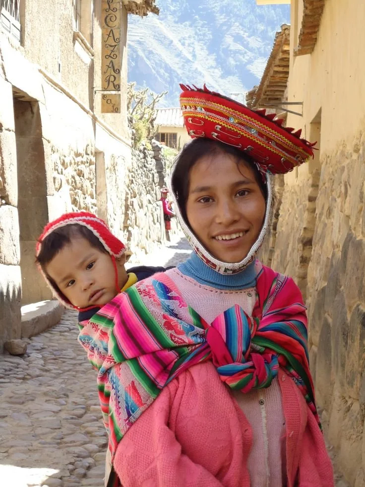 Along a cobbled village street nestled in the Andean highlands, a young mother beams with warmth and quiet pride. Her baby rests against her back, bundled snugly in a vivid striped manta tied across her chest in a firm knot—bright pinks, greens, and
