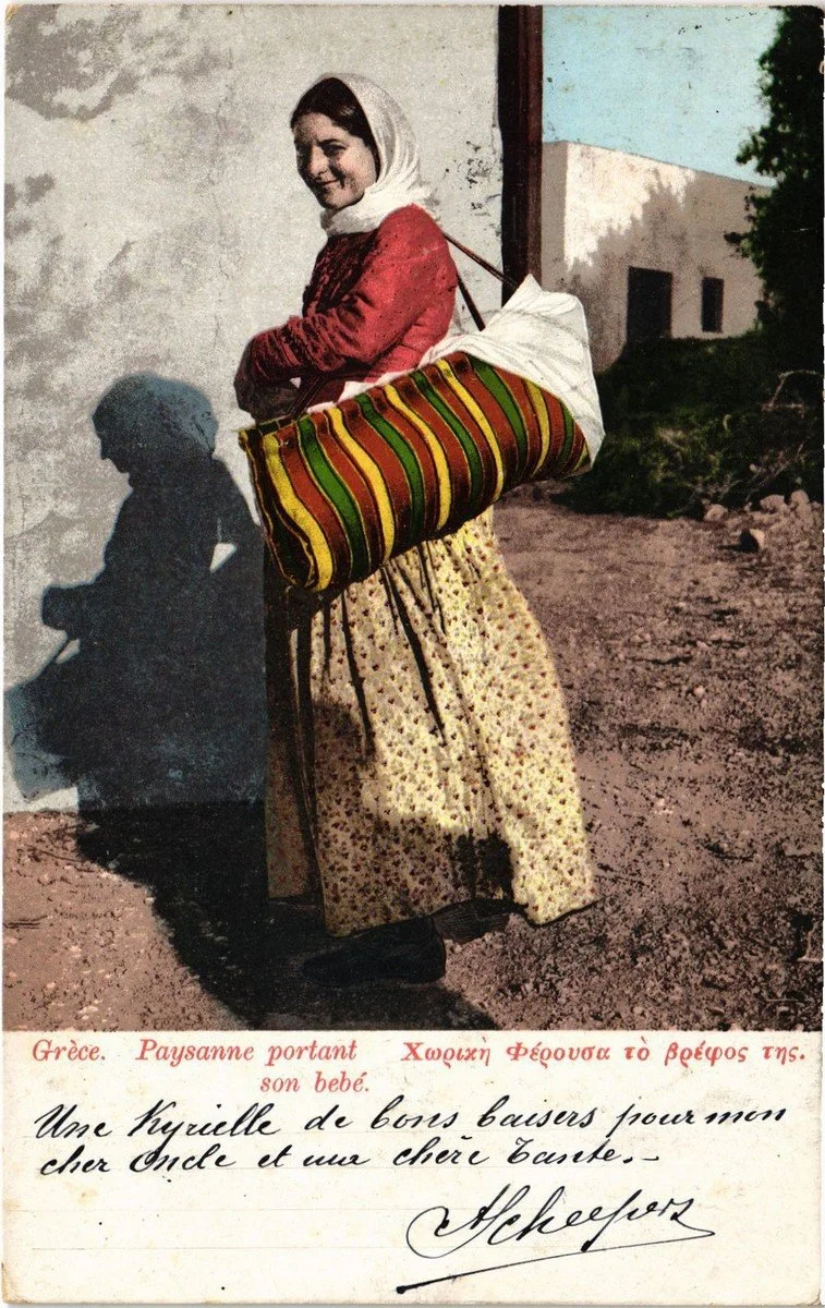 In this colourised early 20th-century postcard from Greece, a rural mother carries her baby in a vividly striped cradle bag — slung across her body and secured over her shoulder by wide leather straps. The bag is long and padded, likely stuffed with