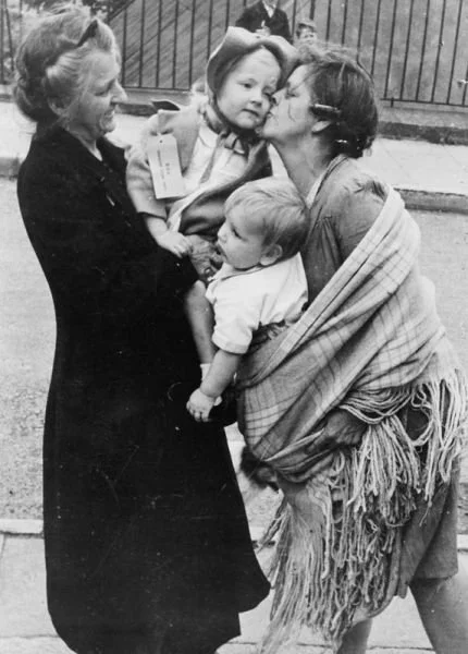 In wartime Britain, a mother kisses her daughter goodbye as the child prepares to be evacuated to safety in the countryside. Her younger sibling is carried in a traditional shawl, wrapped snugly across the mother’s body and supported on her hip. The